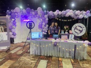 The front office manager, Aleta and marketing coordinator, Natalie are standing in fron tof the Andrews Braces booth at Divas' Day Out Reno 2024. There are 2 purple baloon arches, a photo booth and a raffle prize table