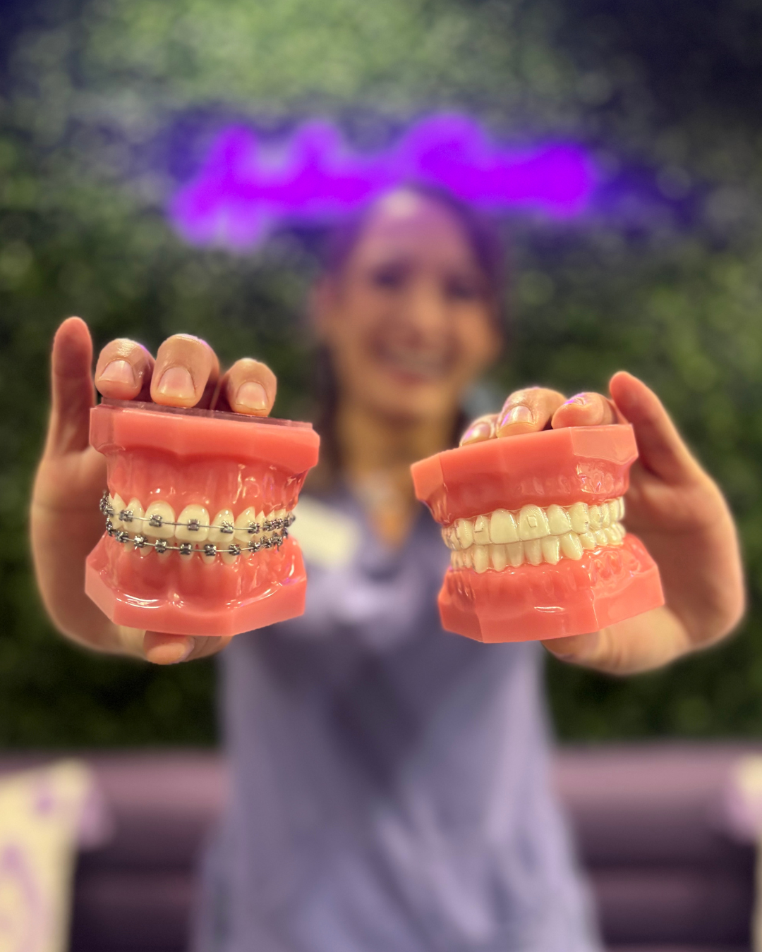 Megan, our treatment coordinator, is holding up two typodonts, one with braces on, and one with clear aligners on